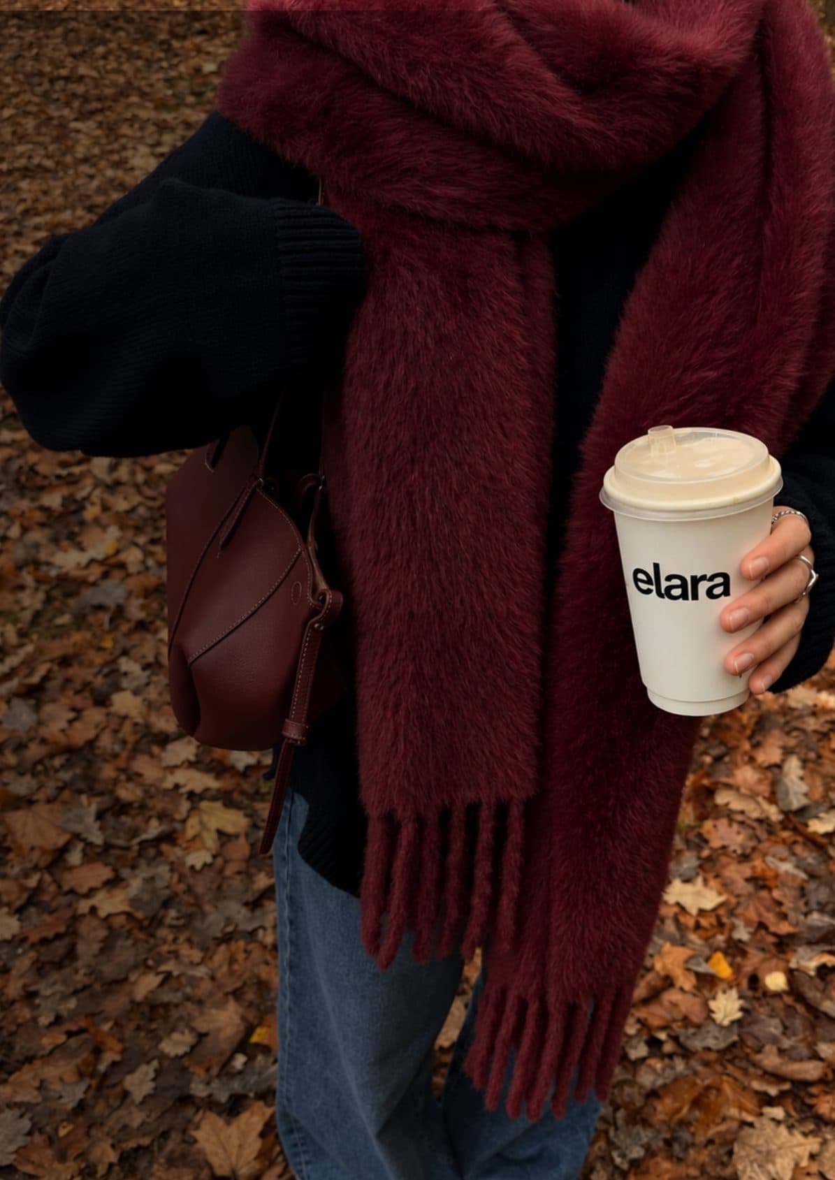 A woman in a burgundy scarf holding an Elara coffee cup over autumn leaves