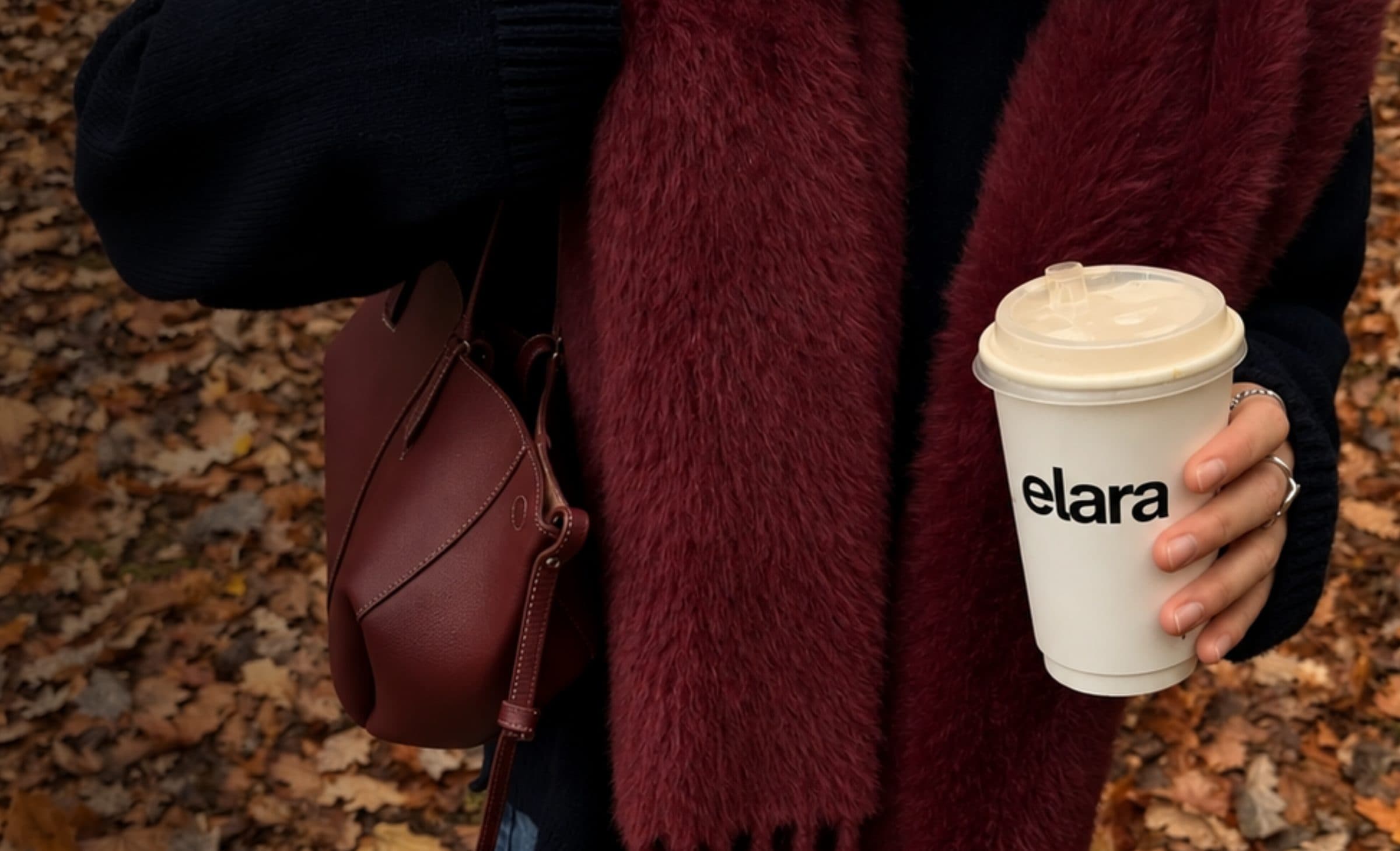 A woman in a burgundy scarf holding an Elara coffee cup over autumn leaves